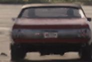 A dark red coupe pulling away across the forecourt, its tail lights running the full width